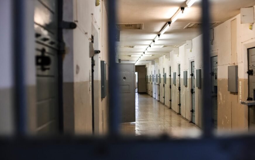 A long prison corridor with rows of metal doors viewed through dark bars